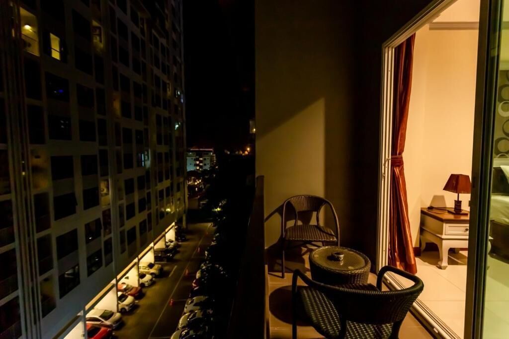 A balcony with a view of parked cars and buildings, a small table with chairs, and a night-time setting.