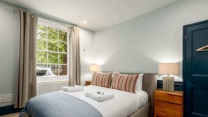 Un lit bien fait, recouvert d’une couverture bleue, une table de chevet avec une lampe et une fenêtre avec des rideaux.