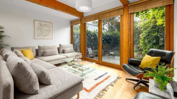 Un salon moderne avec un canapé d’angle, une table basse en verre et un fauteuil en cuir noir. De grandes fenêtres offrent une vue sur la verdure environnante.