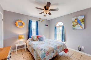 Quarto com cama, ventoinha no teto, mesa de cabeceira, abajur e janela com cortinas.