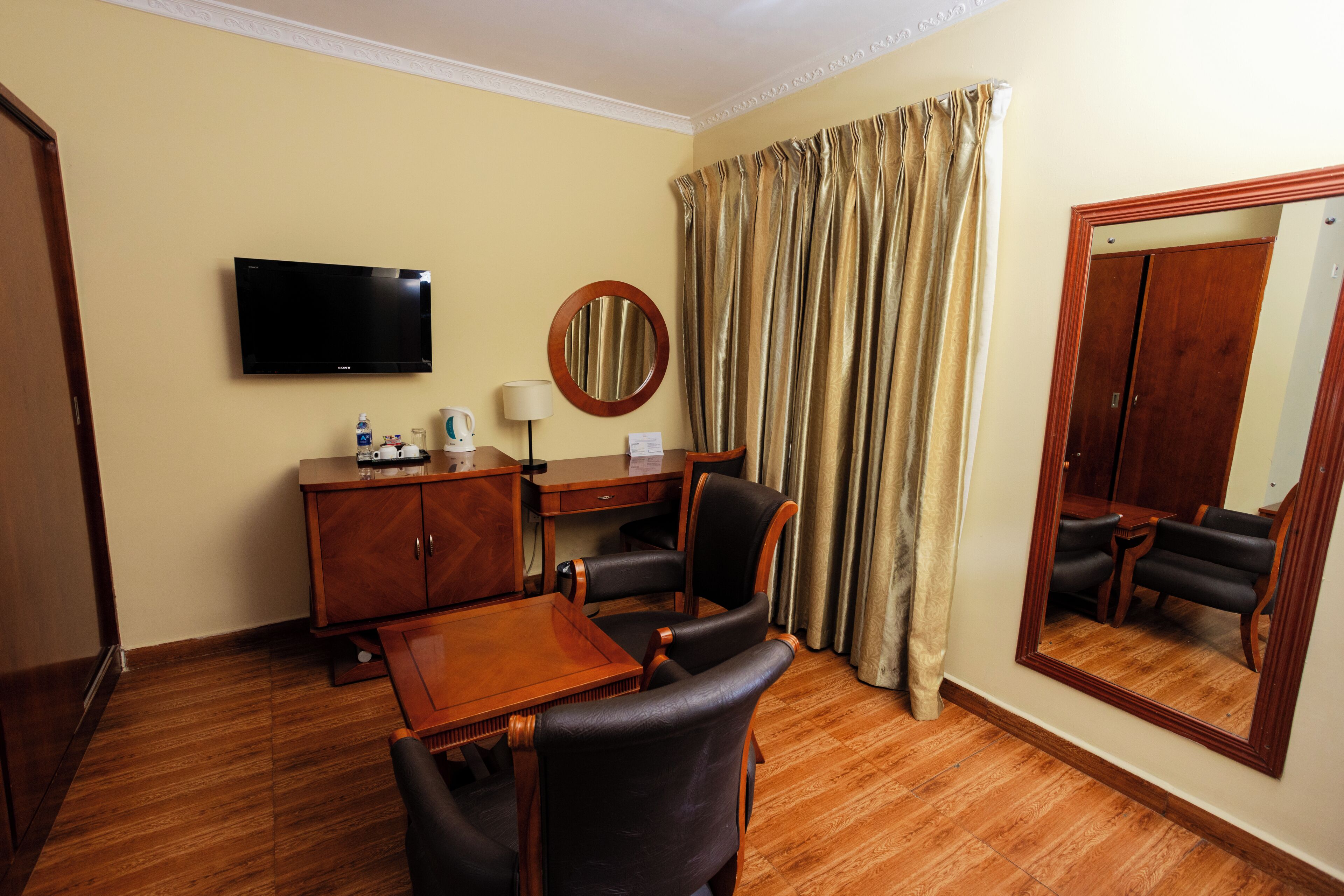 A hotel room with a flat-screen TV, a wooden desk, a chair, a mirror, and a large window with curtains.