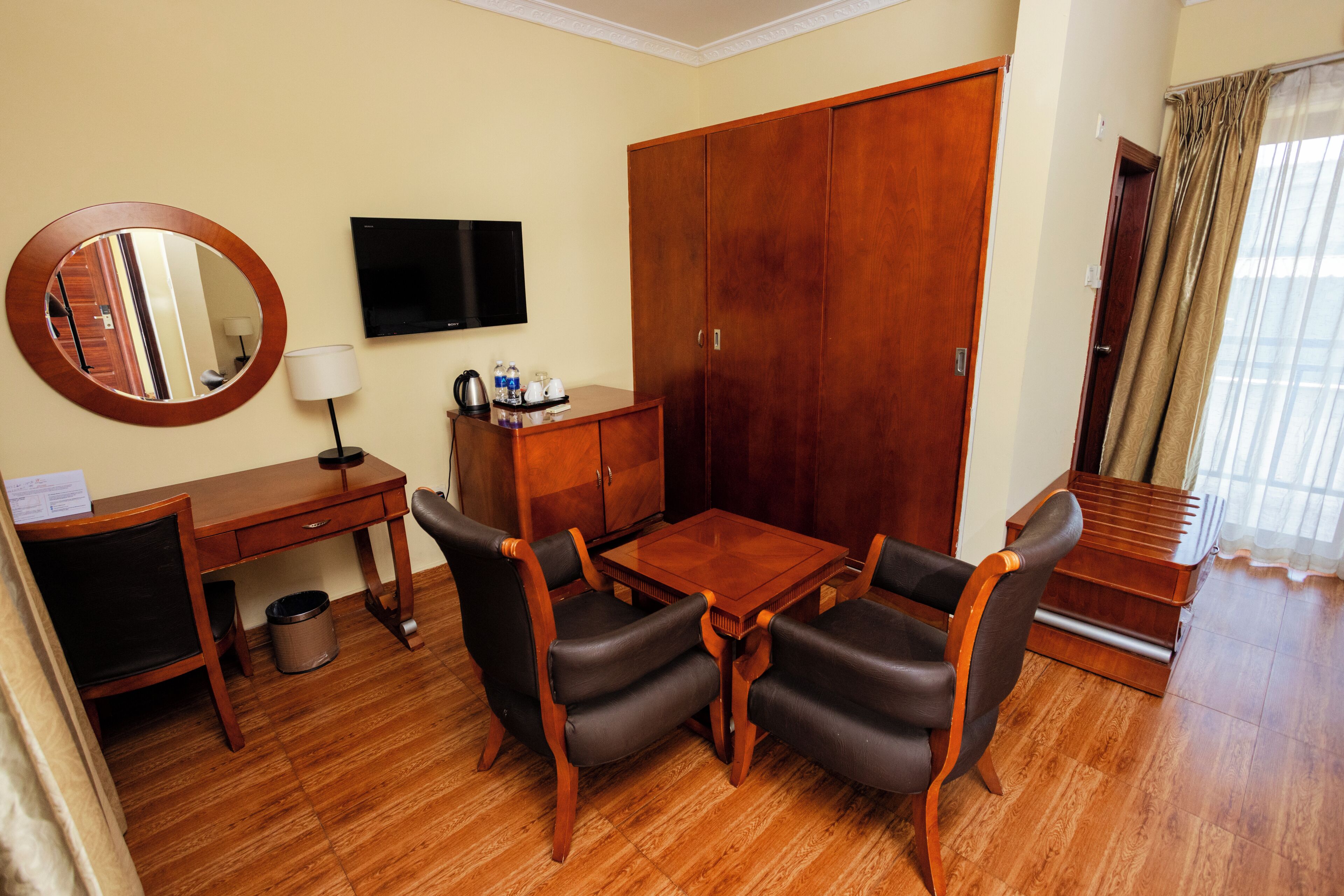 A hotel room with a wooden desk, two chairs, a TV, a wardrobe, and a small table with a lamp.