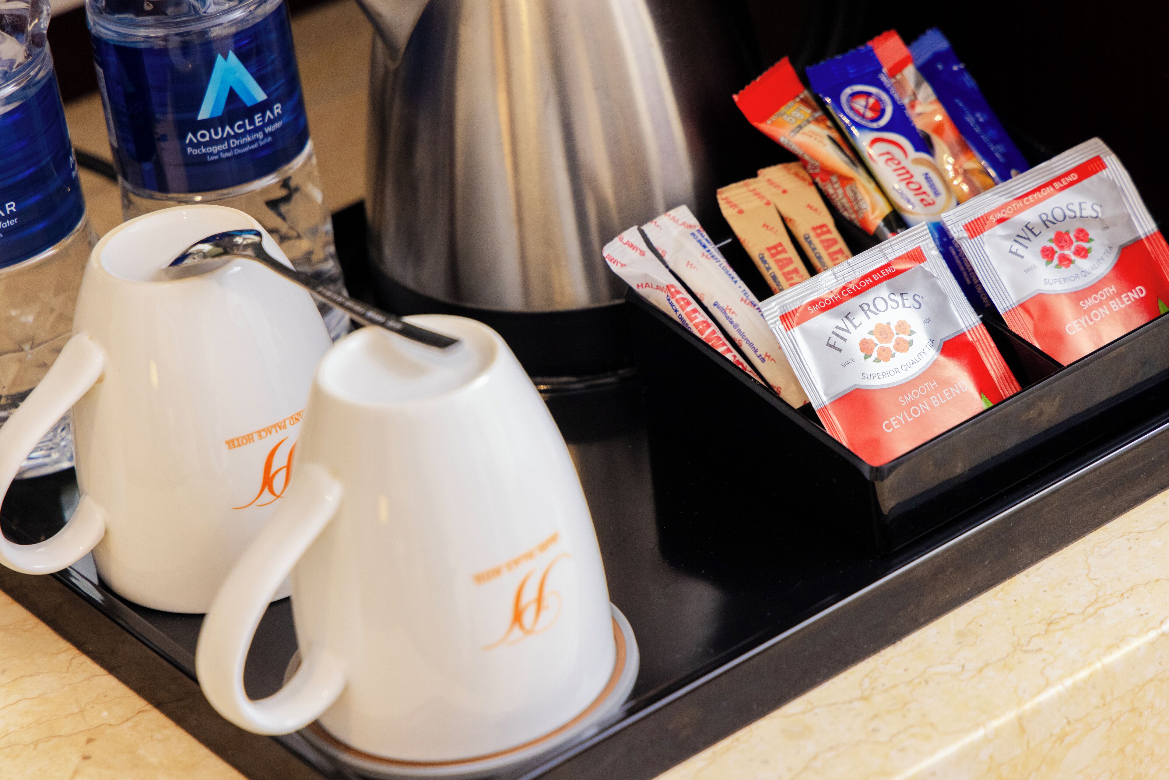 Two white coffee mugs with orange logos, a stainless steel kettle, and a tray with tea packets and a coffee machine.