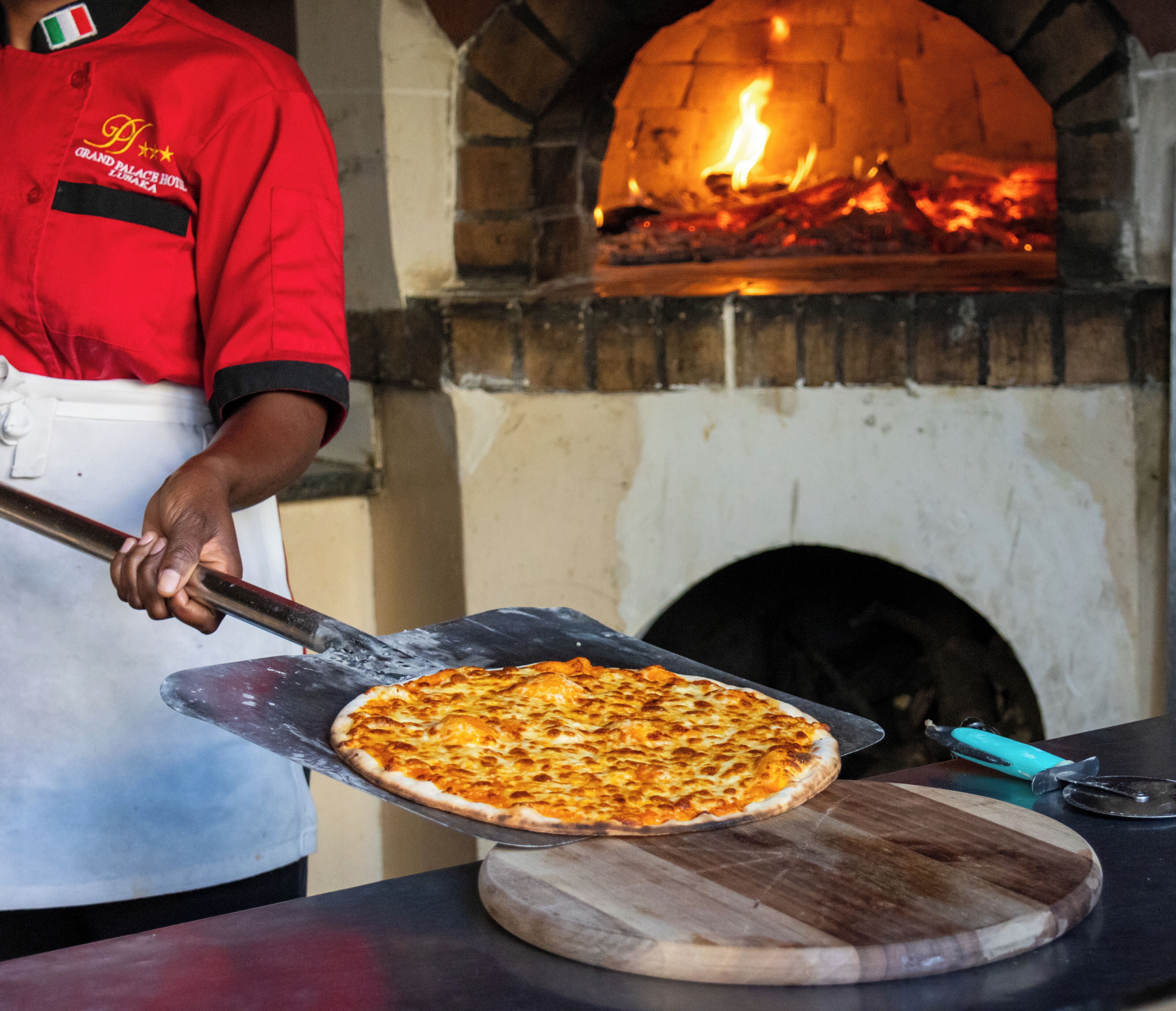 A pizza being served from a brick oven, with a chef in a red uniform.