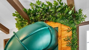 A green spiral slide in a room with wooden flooring, a door, and a wall with a wooden door and greenery. - Disney Explorer - Sauna, Theater, Arcade and More! (Garden Grove)