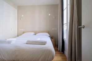 A neatly made bed with white linens and a folded towel, a bedside table with a lamp, and a window with curtains. - Charming Parisian 1BDR Apt Near Gare du Nord (Paris)