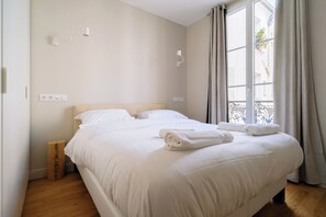 A hotel room with a large bed, a bedside table, a wooden stool, and a window with curtains. - Charming Parisian 1BDR Apt Near Gare du Nord (Paris)