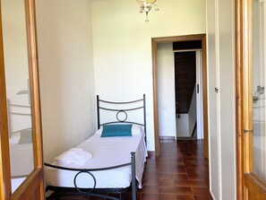 Ein Einzelbett mit Metallgestell, weißer Bettwäsche und einem türkisblauen Kopfkissen in einem schmalen Raum mit gefliestem Boden und einem Holztürrahmen.