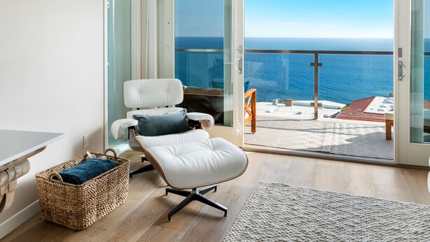 A balcony with a view of the ocean, a white chair, a wicker basket, and a wooden floor.