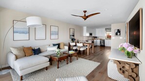 A modern living room with a sofa, coffee table, and a dining area with chairs. There is a ceiling fan, artwork on the walls, and a kitchen in the background.