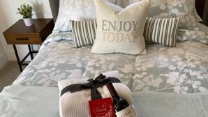 A neatly made bed with floral bedding, two pillows with the text "ENJOY TODAY," a bottle of wine, and a box of Lindor chocolates.