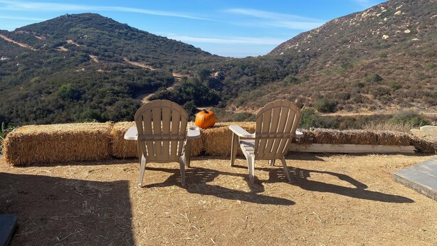 Two wooden chairs facing a scenic view of a valley with a large pumpkin on a hay bale.