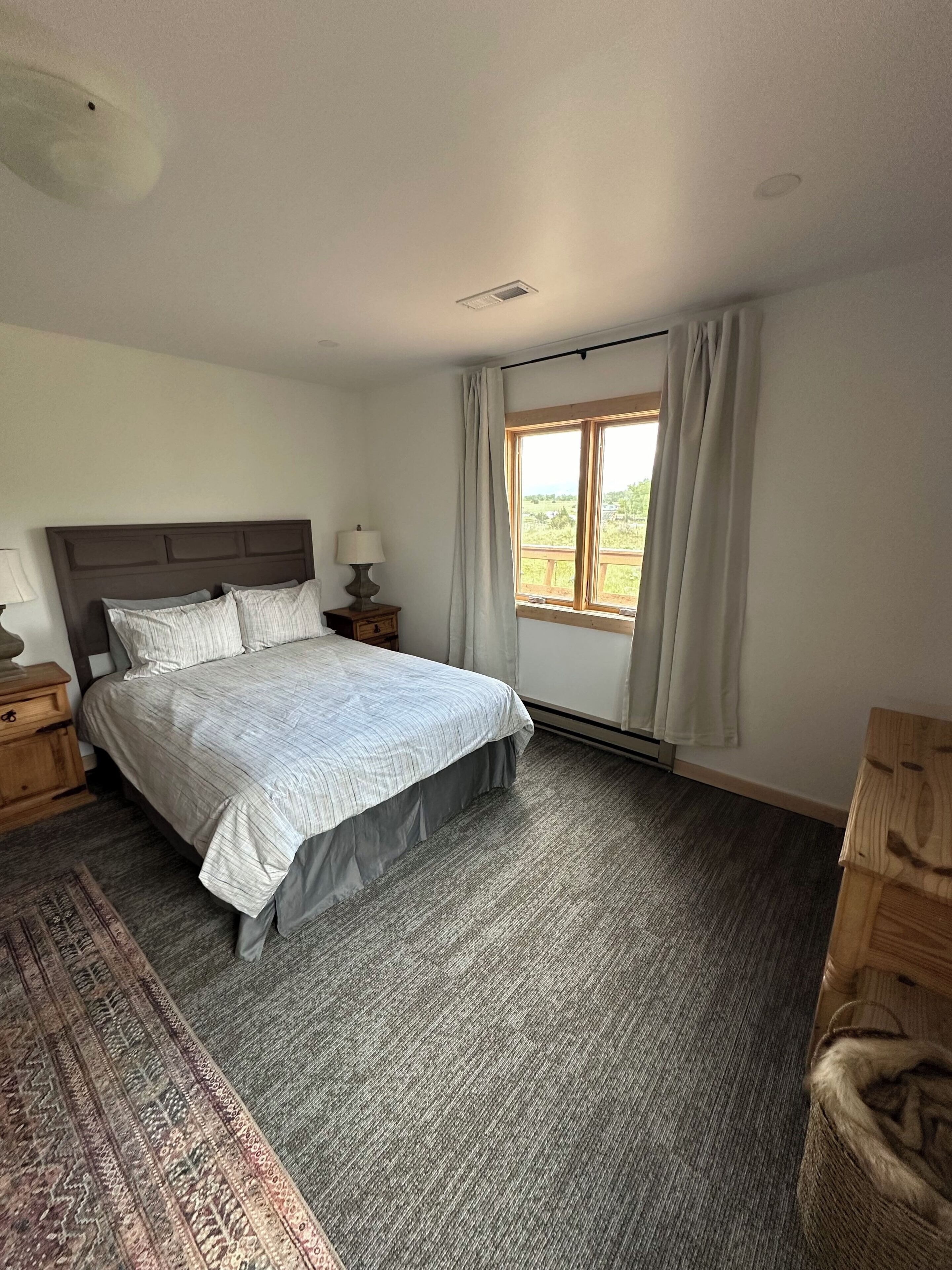 A bedroom with a bed, nightstands, a lamp, a window with curtains, and a view of greenery outside.