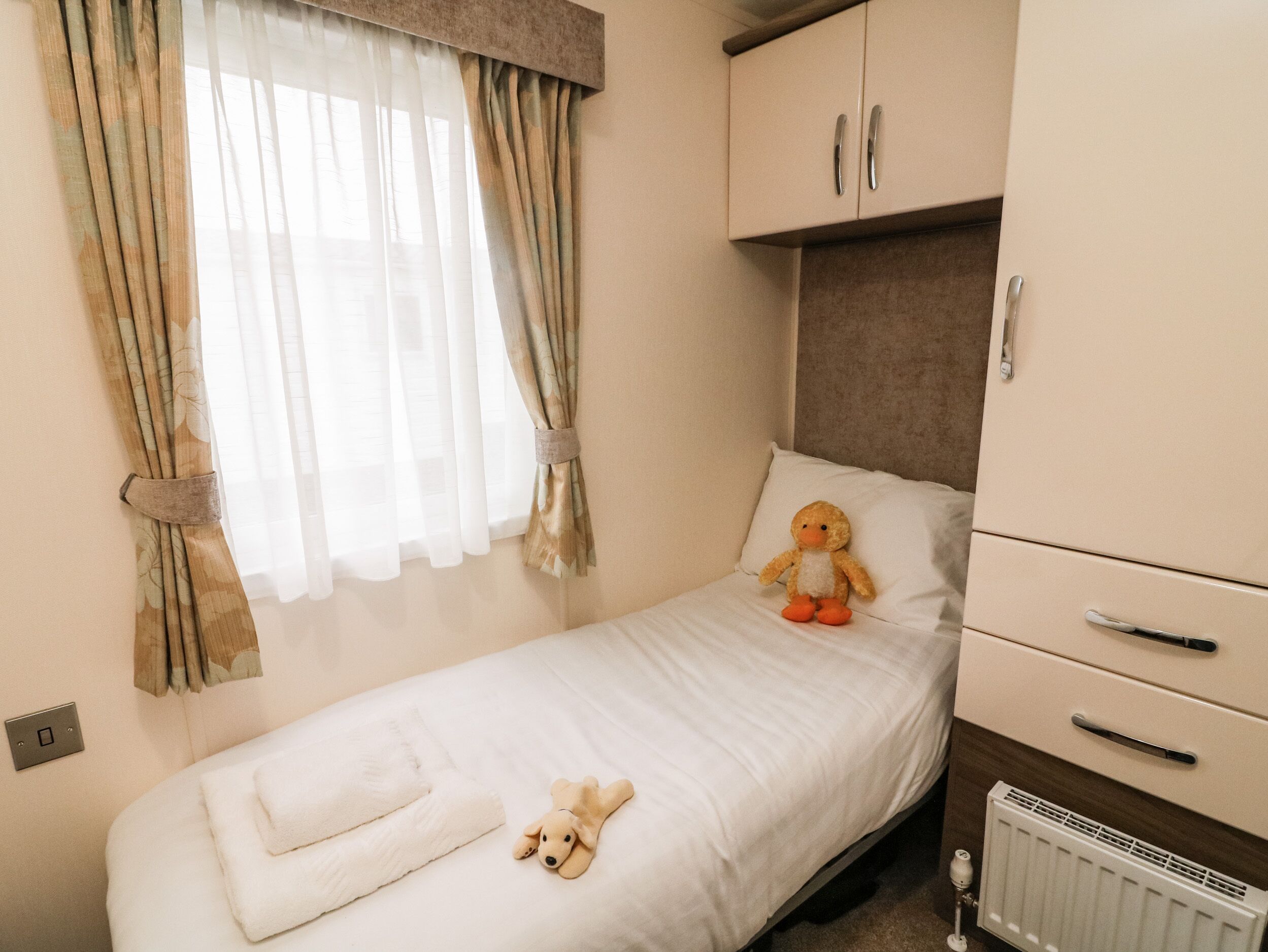 Un lit simple avec une couette blanche, un ours en peluche et un jouet en peluche représentant un chien posés sur le lit. Il y a un radiateur sous le lit et une fenêtre avec des rideaux.