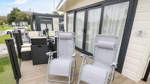 A deck with two white canvas chairs, a wicker table, and a glass door leading to a covered area. - 3 Yealands (Carnforth)