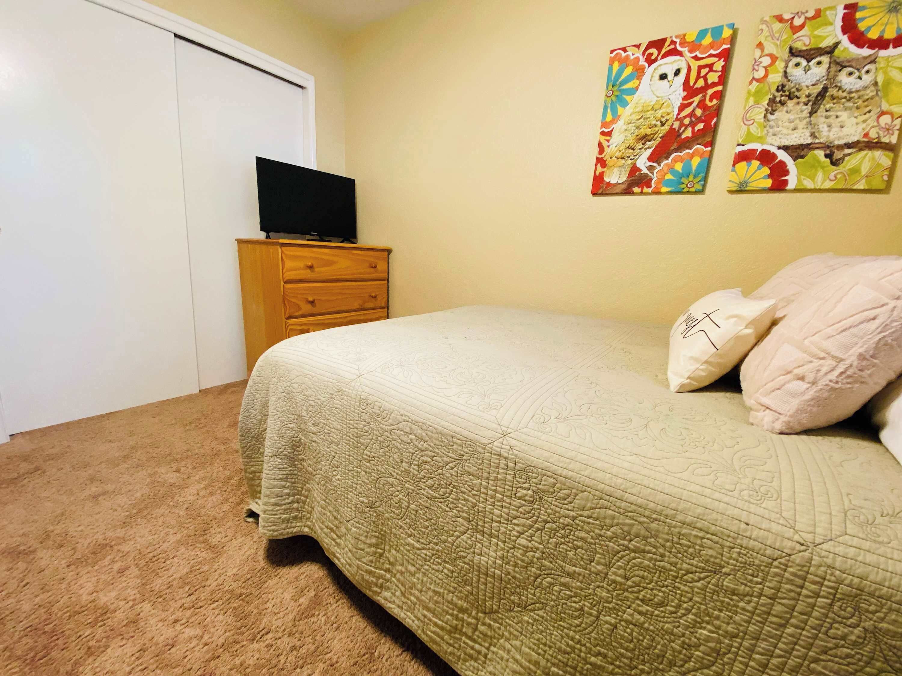 A bedroom with a bed, a wooden dresser, a television on top, and two wall-mounted paintings.