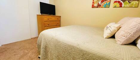 A bedroom with a bed, a wooden dresser, a television on top, and two wall-mounted paintings.