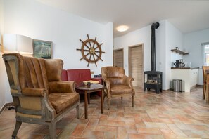 A living room with two leather armchairs, a red sofa, a wooden table, and a ship's wheel decoration. A black stove is in the corner.