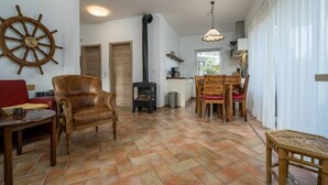 Un espacio de sala acogedor con un sillón de cuero, una mesa de madera y una mesa de comedor con sillas. Un adorno con timón de barco está en la pared, y una estufa de leña al fondo.