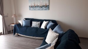 Un salon avec un canapé bleu foncé, de nombreux coussins, un tableau représentant des voiliers, une lampe et une fenêtre avec des rideaux.