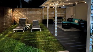 A covered outdoor seating area with lounge chairs and a sofa, surrounded by a wooden fence and a grassy lawn.