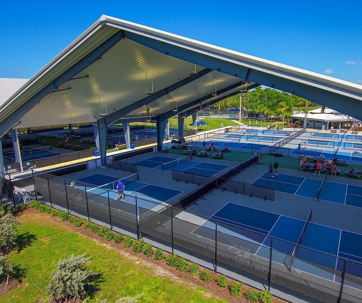 Ein Tennisplatz mit mehreren Spielfeldern, einer überdachten Zuschauertribüne und einer weitläufigen Grünfläche mit Bäumen und einem Gebäude im Hintergrund.