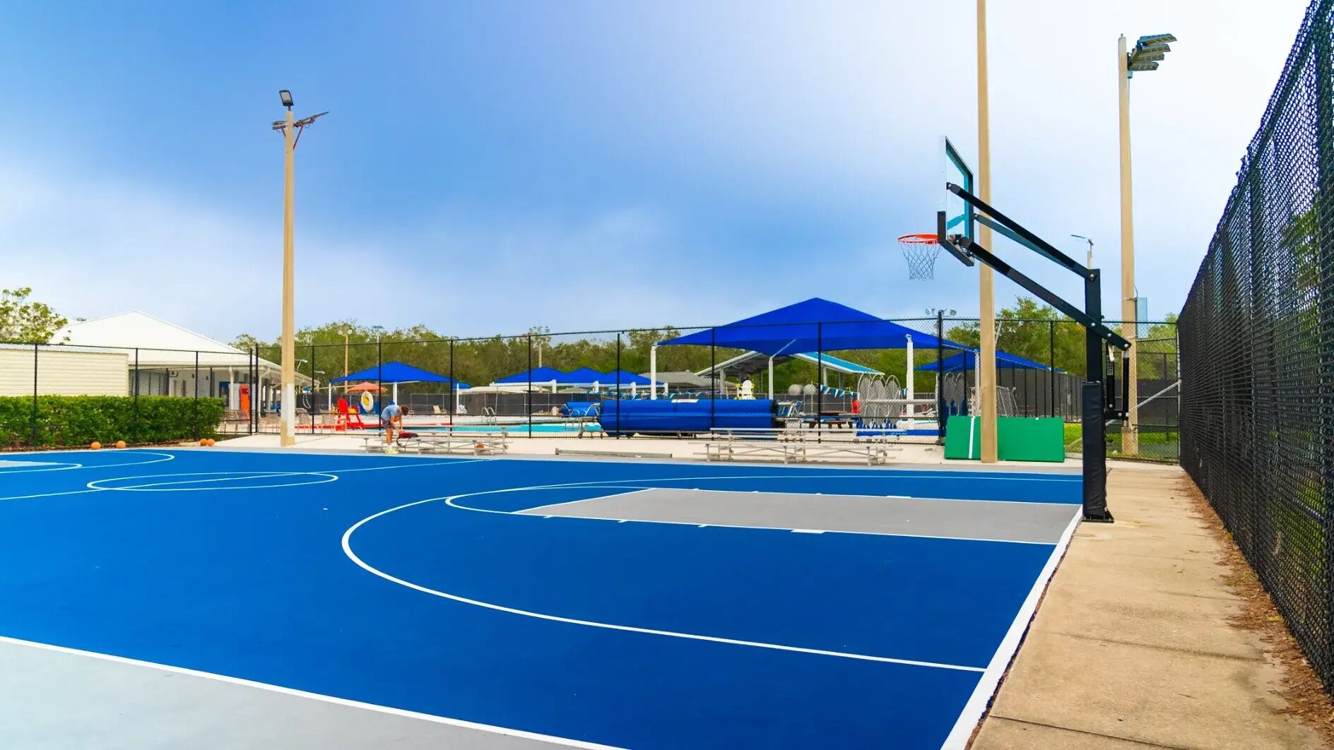 An outdoor basketball court with blue flooring, surrounded by a fence, and shaded by blue umbrellas.