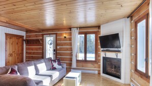 A log cabin interior with a sofa, television, fireplace, and large windows.