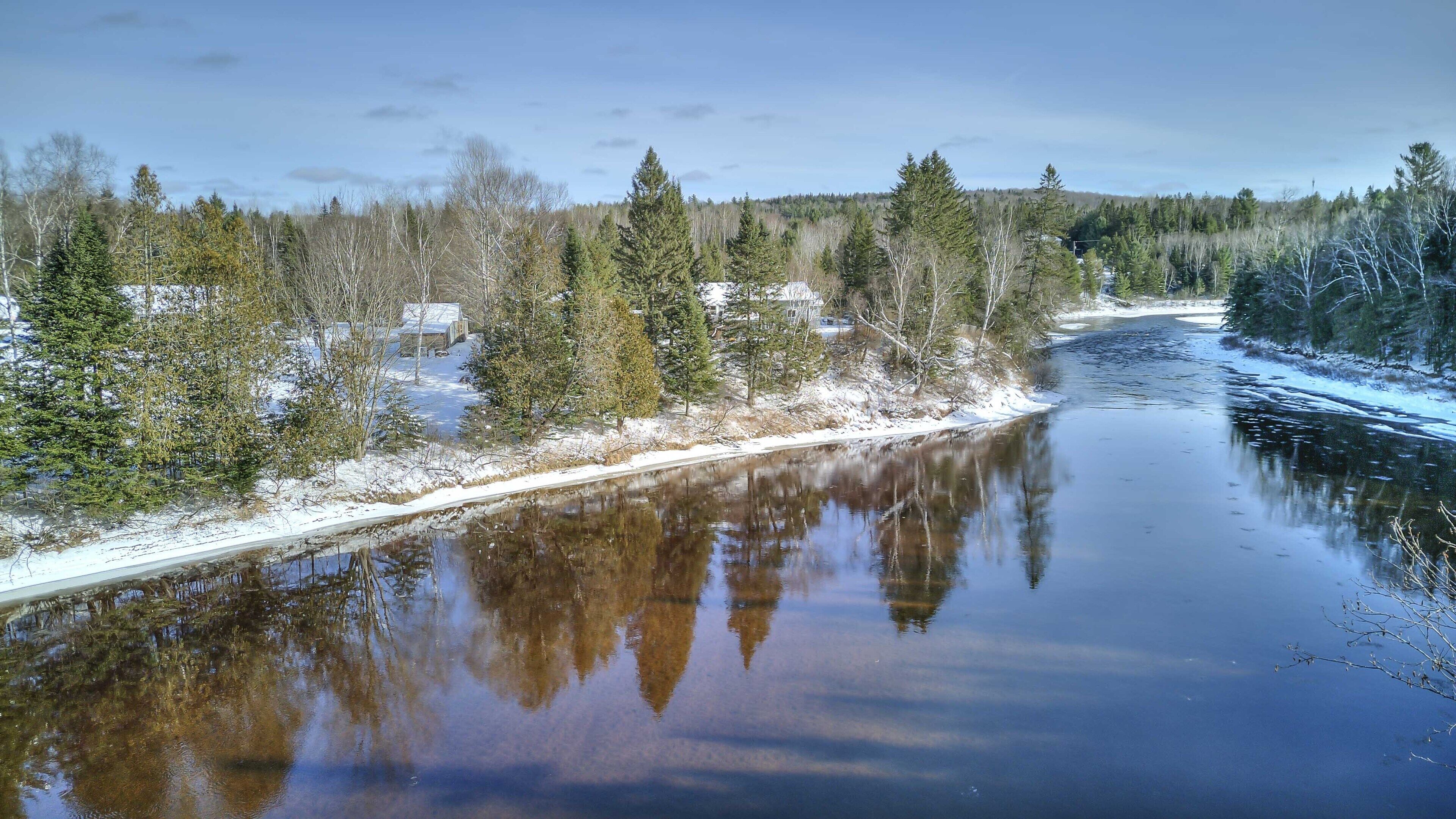 A river with reflections of trees and buildings, surrounded by snow-covered banks and a forested area.
