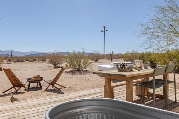 A wooden deck with a table and chairs, a metal container, and a desert landscape with sparse vegetation and distant mountains.
