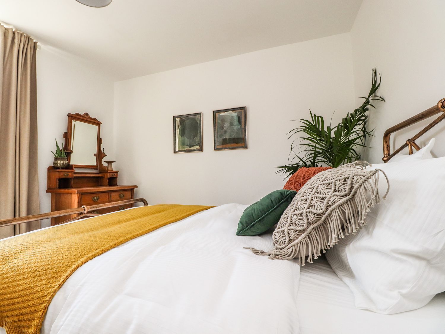 A bedroom with a bed, a wooden dresser, a mirror, two framed pictures, and a potted plant.