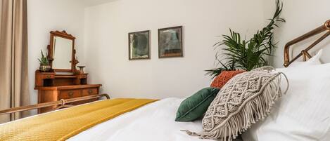 A bedroom with a bed, a wooden dresser, a mirror, two framed pictures, and a potted plant.