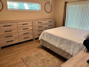Une chambre à coucher avec un lit, une commode en bois et une fenêtre avec des stores.