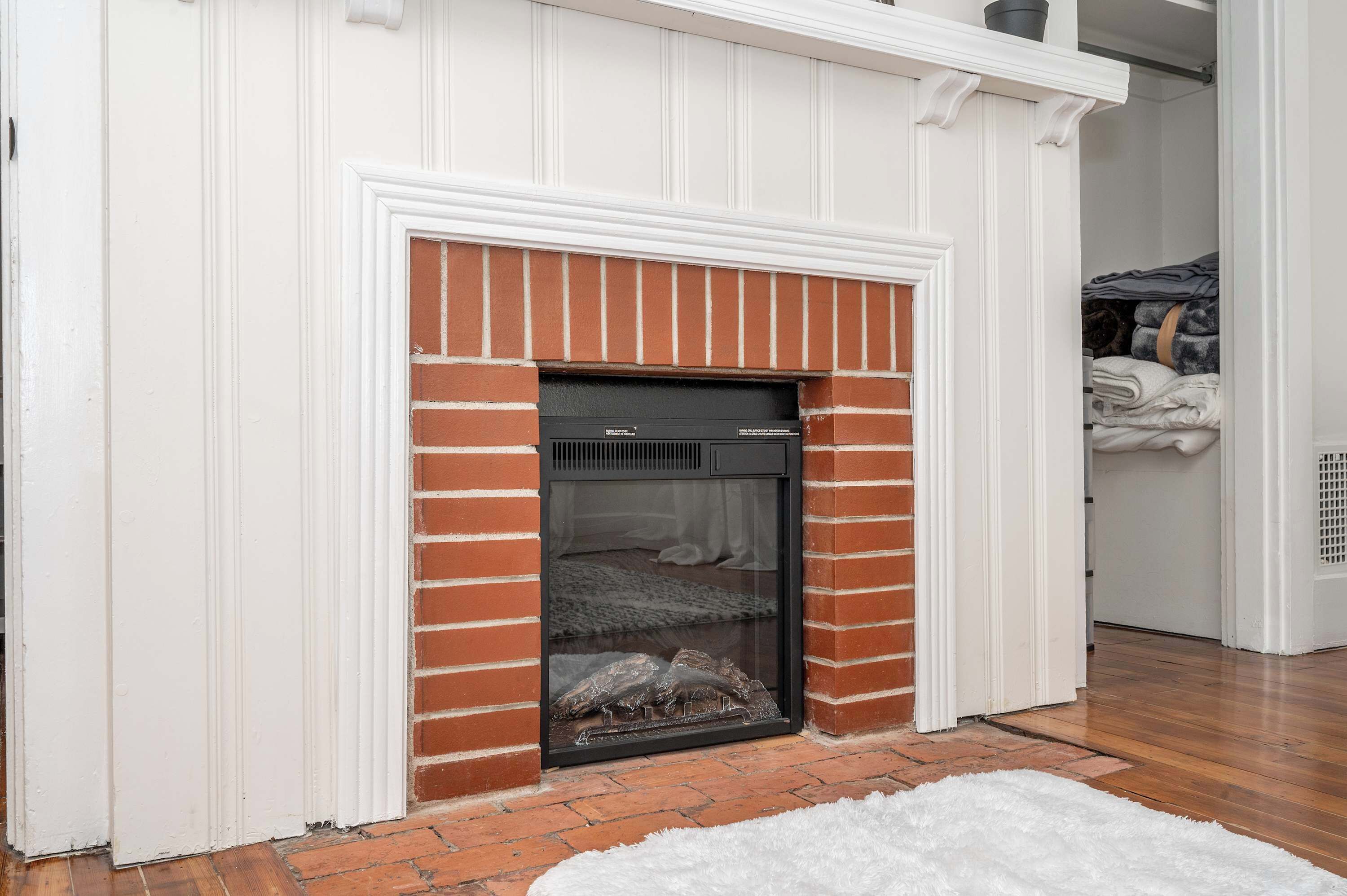 A fireplace with a brick surround and white trim, located in a room with wooden flooring and a white rug.