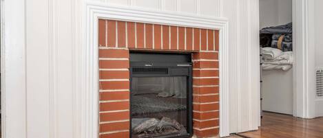 A fireplace with a brick surround and white trim, located in a room with wooden flooring and a white rug.