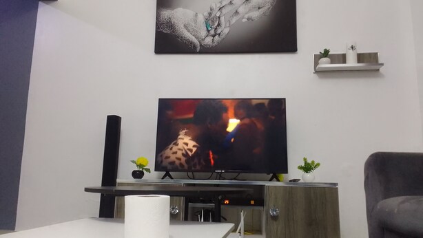 Una sala de estar con una televisión de pantalla plana, una mesa blanca con un rollo de papel higiénico, un sillón gris, un mueble de madera y un estante pequeño con objetos decorativos.