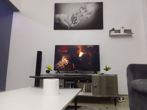 A living room with a flat-screen TV, a white table with a roll of toilet paper, a grey armchair, a wooden cabinet, and a small shelf with decorative items.