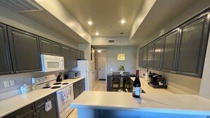Une cuisine moderne avec des meubles de couleur sombre, un plan de travail blanc, un four encastré, un four à micro-ondes et un réfrigérateur. On y trouve un coin repas avec une table et des chaises, ainsi qu’une bouteille de vin posée sur le plan de travail.