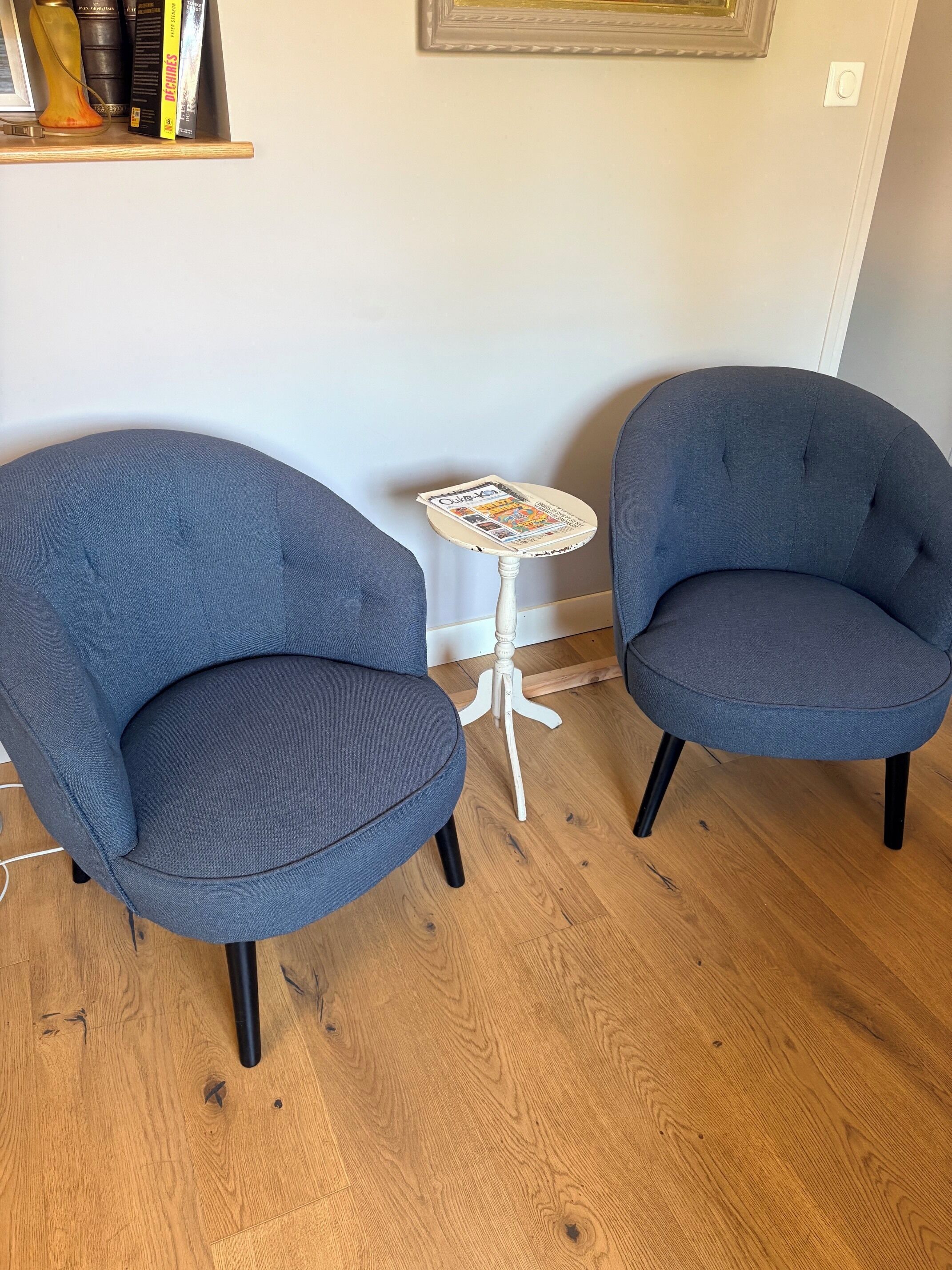 Dos sillones azules, una mesita pequeña con superficie estampada y una estantería con libros y objetos decorativos.