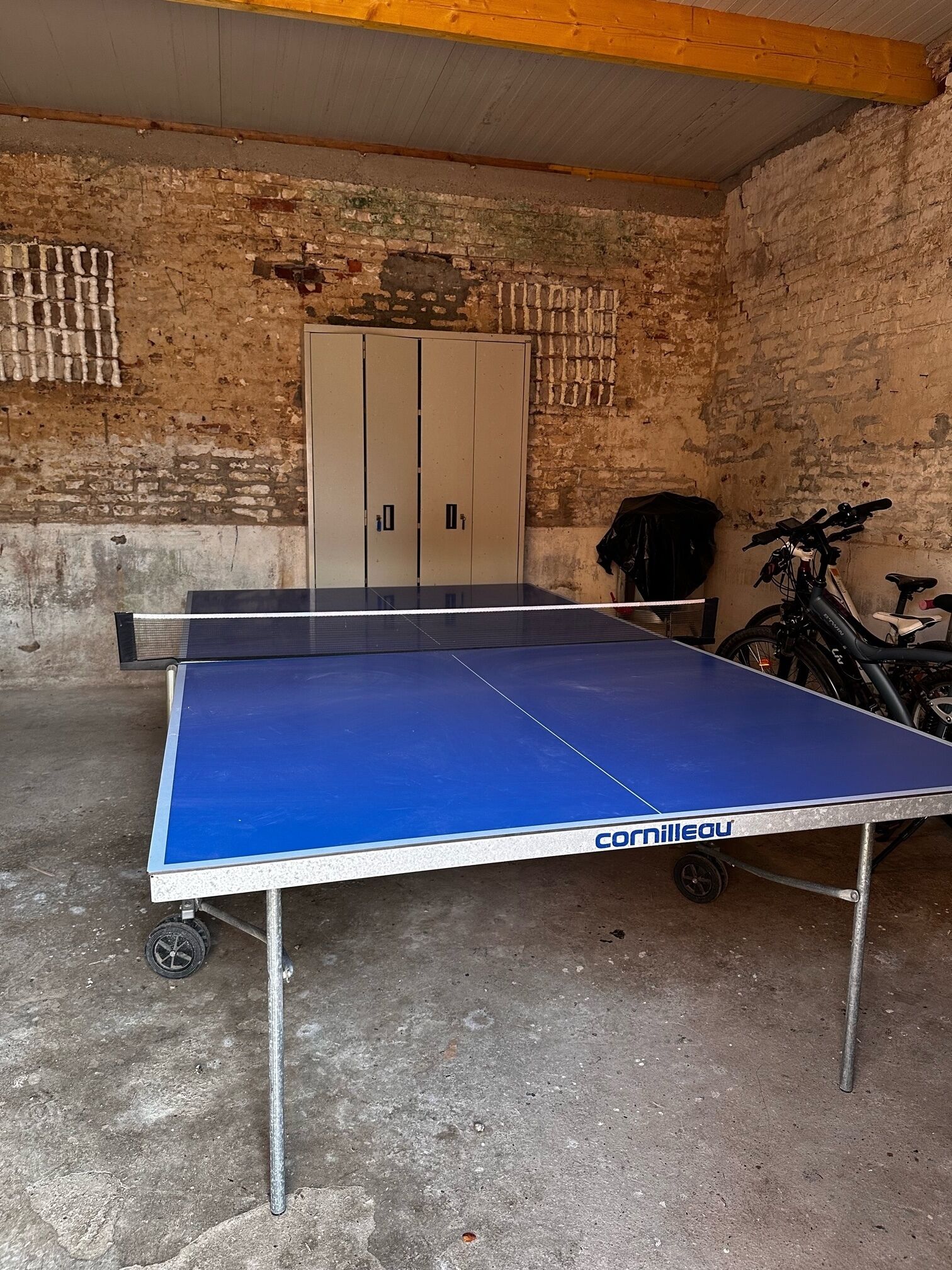 Una sala con pavimento in cemento e pareti in mattoni, con un tavolo da ping pong su ruote, una bicicletta e un armadio.