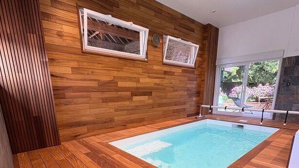 A modern indoor swimming pool with wooden flooring and walls, large windows, and a view of the outdoors.