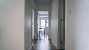 Kapea käytävä, jossa on puulattia ja valkoiset seinät, johtaa huoneeseen, jossa on suuri ikkuna ja pieni pöytä.