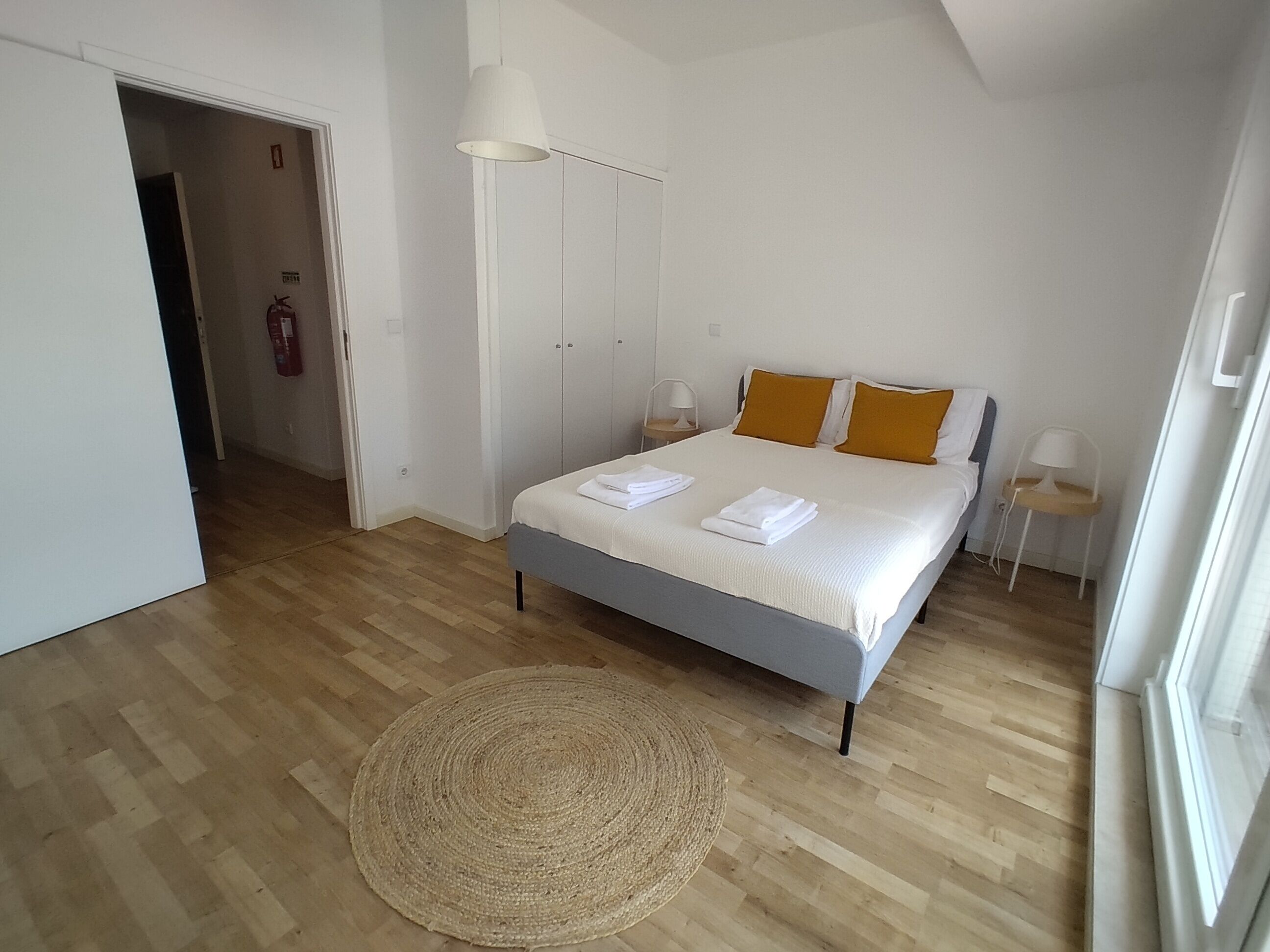 A bedroom with a bed, a bedside table, a lamp, a woven rug, and a door leading to another room.