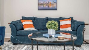 Una sala de estar moderna con un sofá azul oscuro, dos almohadas a rayas naranja y blanco, una mesa de centro de vidrio con un posavasos azul y una fotografía enmarcada de un paisaje urbano en la pared.