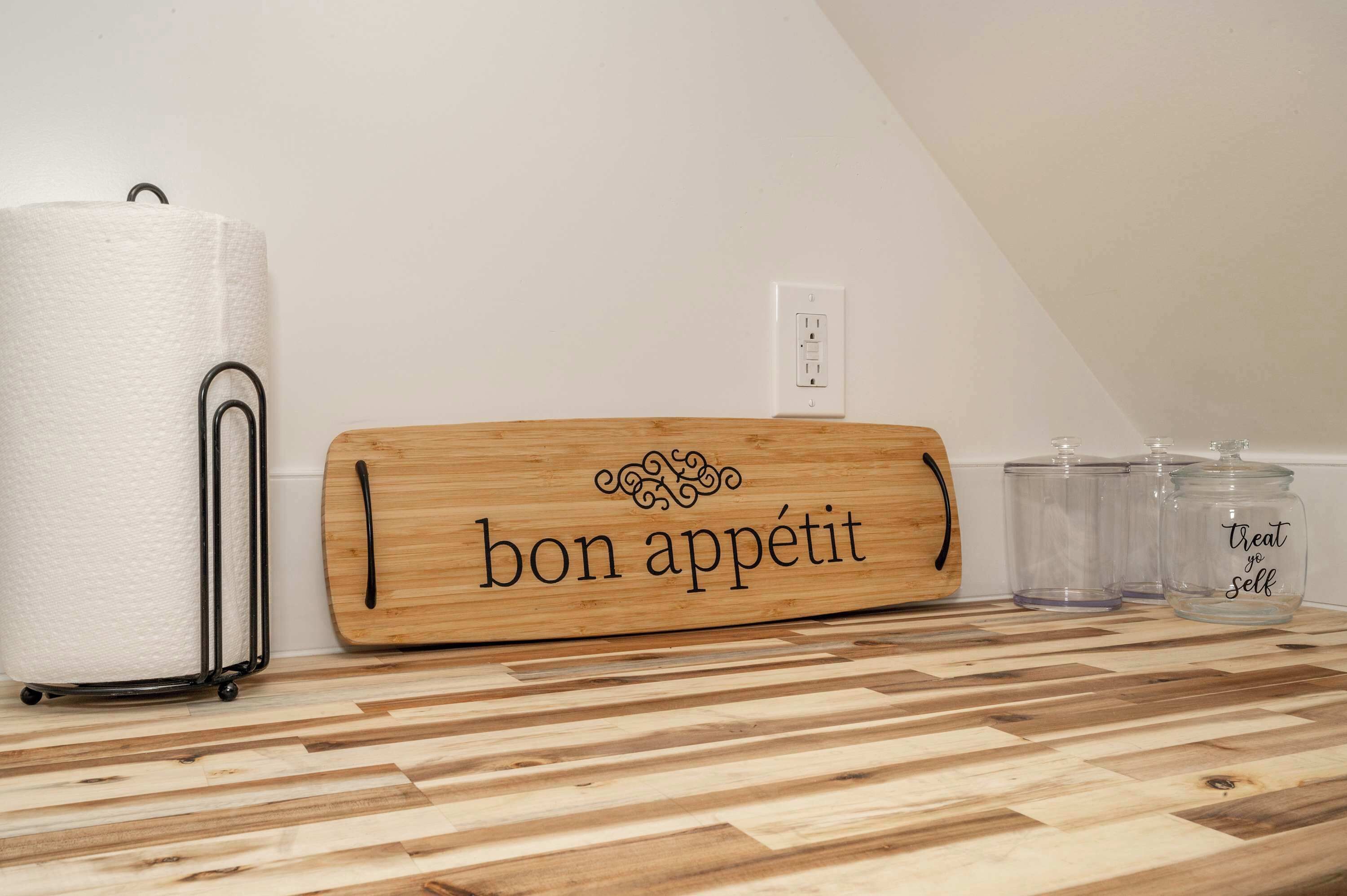 Una encimera de cocina con una tabla de cortar de madera que dice 'bon appétit', un rollo de papel absorbente y dos frascos de vidrio con tapa.