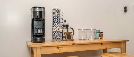 A wooden table with a coffee maker, a French press, and a teapot. There are two stools and a glass jar on the table.