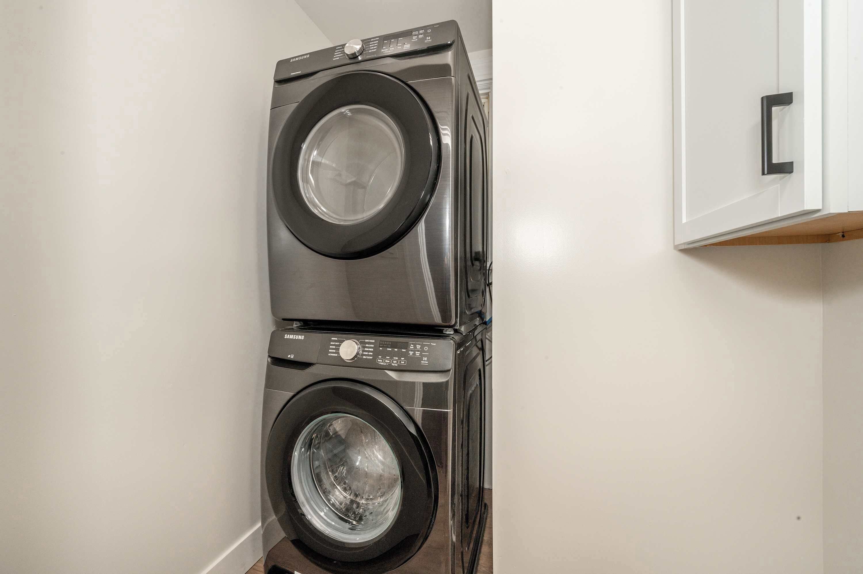 Un appareil combiné lave-linge et sèche-linge, placé dans un coin d’une pièce aux murs blancs et dotée d’un meuble en bois.