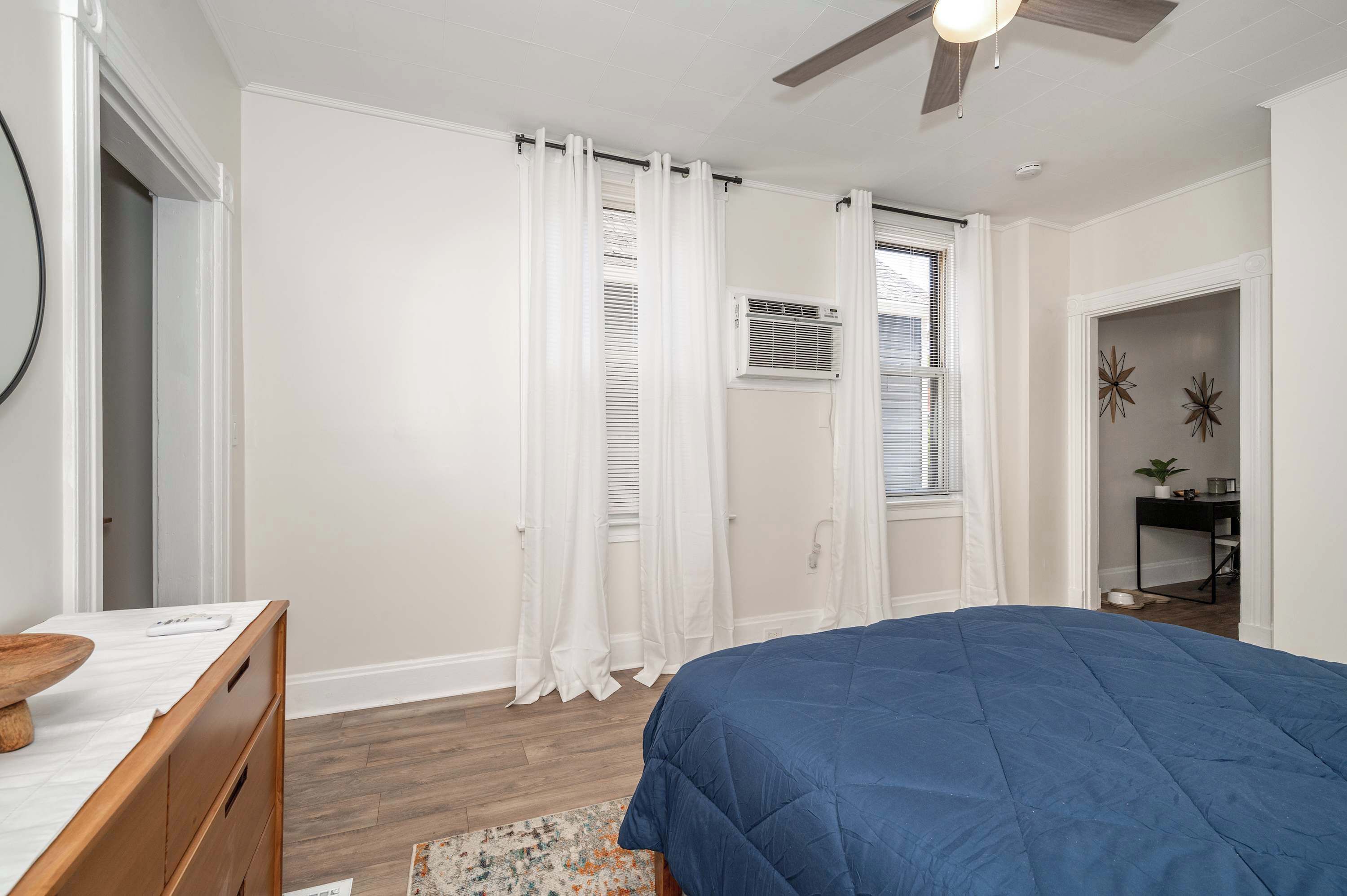 A bedroom with a bed, a desk, and a chair. There is a ceiling fan and a window with white curtains.