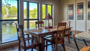 Un comedor con una mesa y sillas de madera, ventanas amplias con vistas a una masa de agua y obras de arte en la pared.