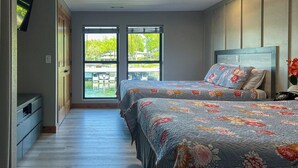 Un dormitorio con dos camas, un televisor montado en la pared y un balcón con vistas a zonas verdes y agua.